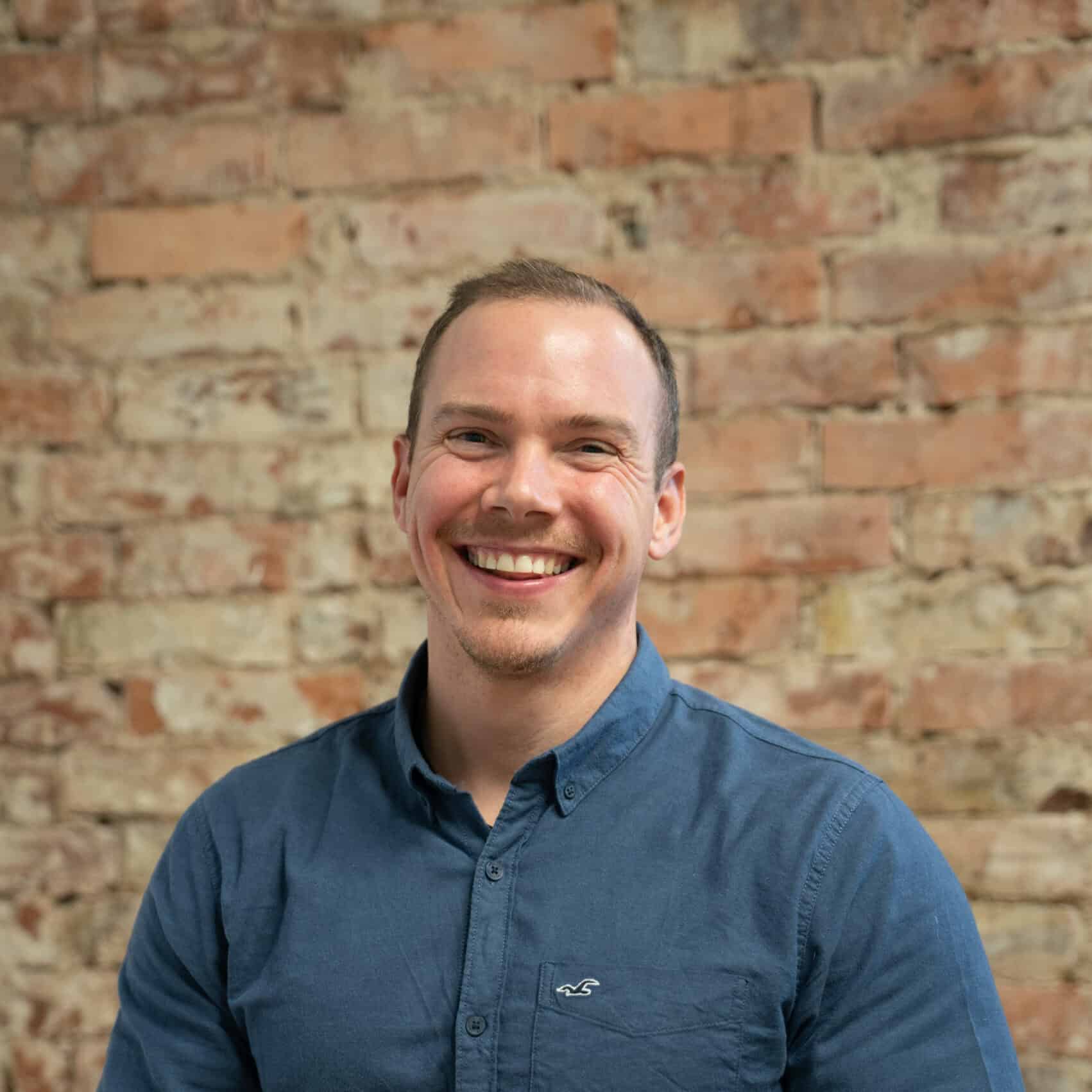 Freundlicher Pflegedienst-Mitarbeiter in blauer Hemd vor Backsteingemäuer, präsentiert professionelle Pflege und Betreuung bei Hahne Pflegedienst, engagiert in der Senioren- und Krankenpflege.